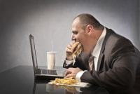 Gruby mężczyzna w garniturze gapi się w laptop jedząc hamburgera, a obok leżą frytki i napój z rurką