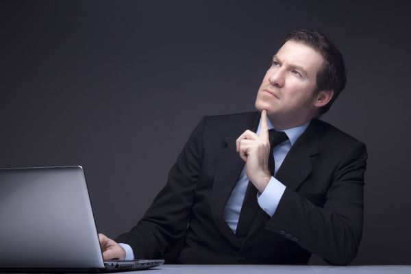 Przystojny mężczyzna w garniturze siedzi przed laptoptem i zastanawia się patrząc w górę i podpierając brodę palcem wskazującym lewej ręki